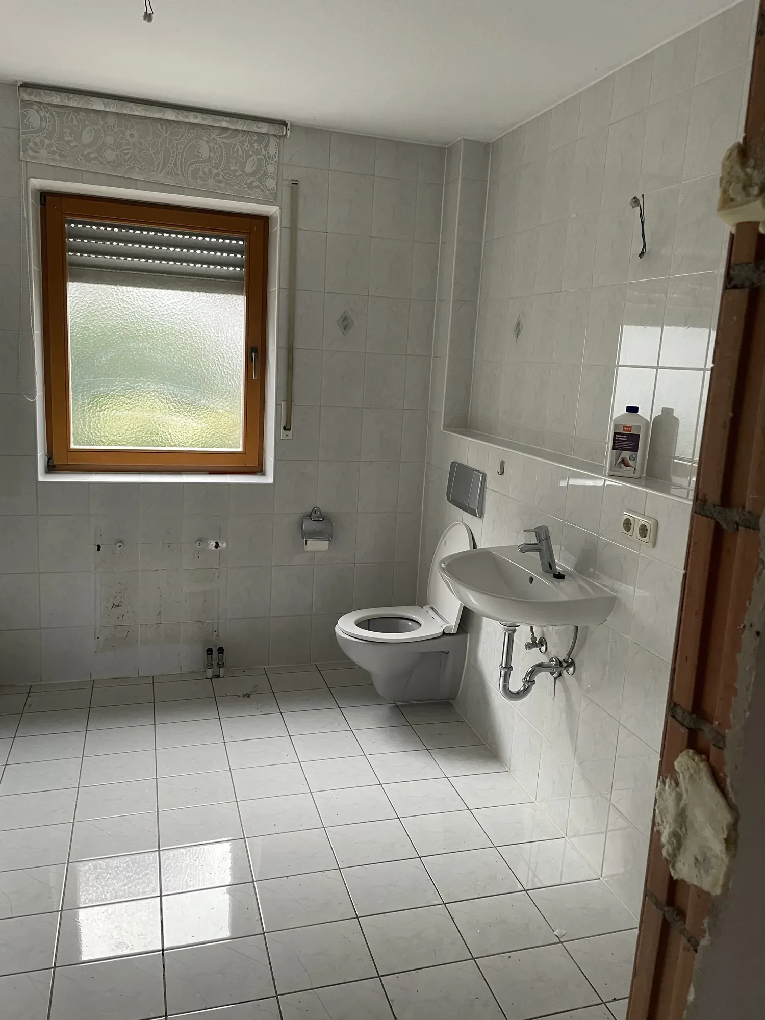 Altes Bad mit weißen Standardfliesen, WC, kleinem Waschbecken und Fenster mit Holzrahmen