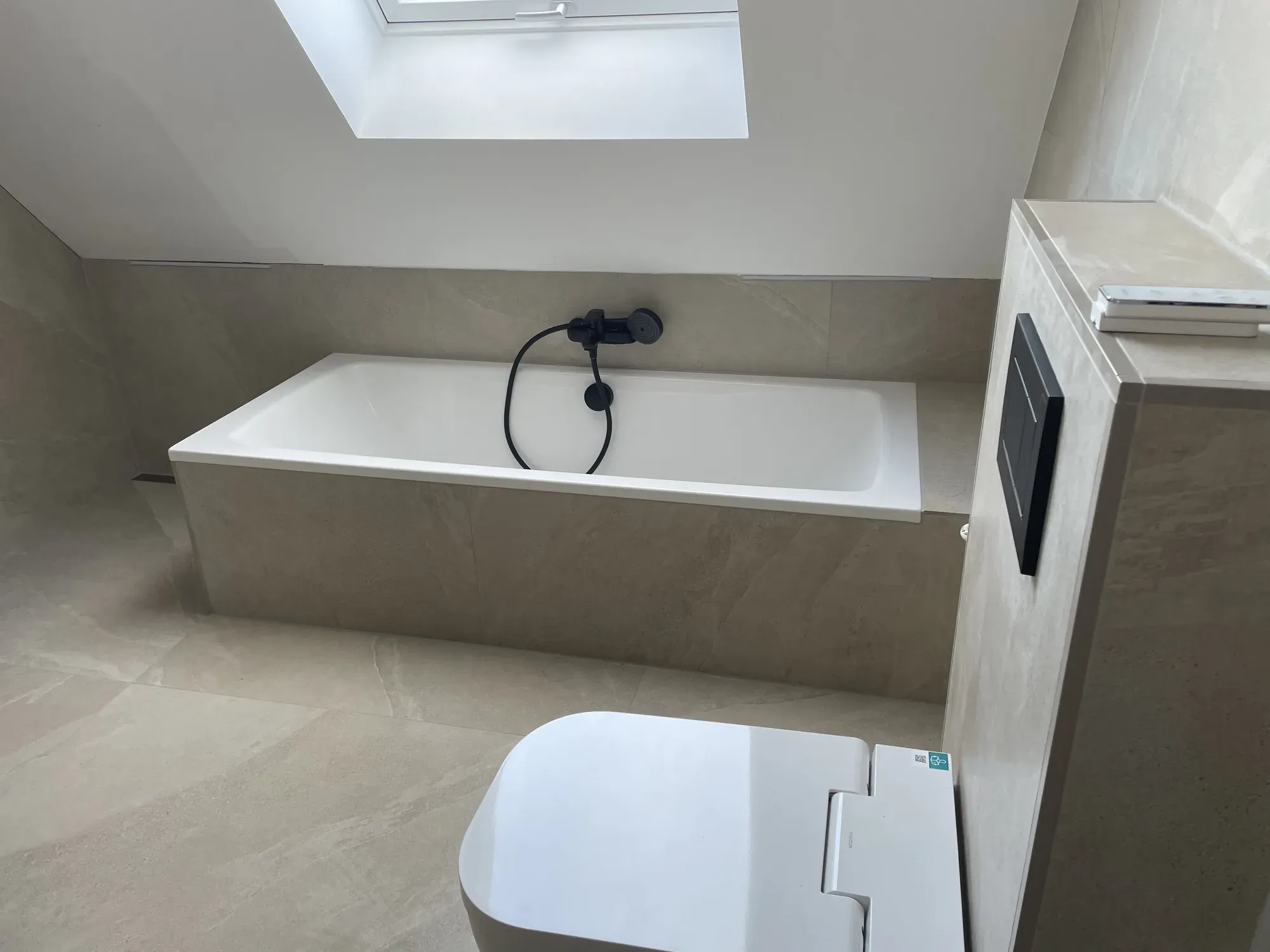 Dasselbe Dachbad nach der Sanierung: neue Wanne unter dem Dachfenster, sandfarbene Fliesen, schwarze Armatur